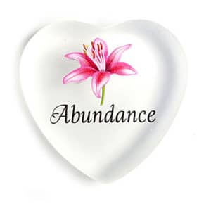 A heart shaped glass paperweight with the word " abundance " and a flower.