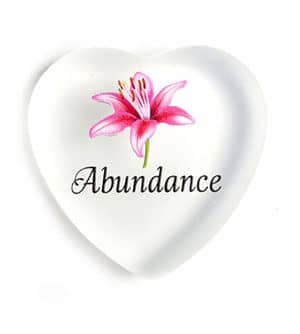 A heart shaped glass paperweight with the word " abundance " and a flower.